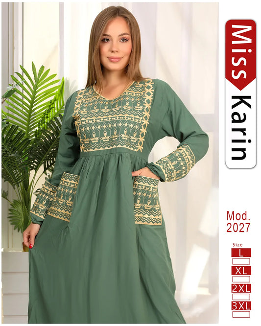 Woman wearing a green dress with gold embroidery, standing indoors with a plant in the background.