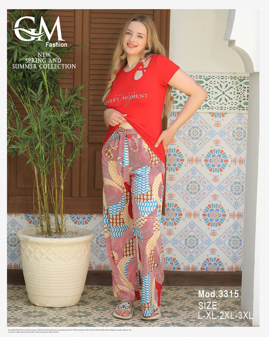 Woman wearing a red top and patterned pants standing next to a plant and decorative wall.
