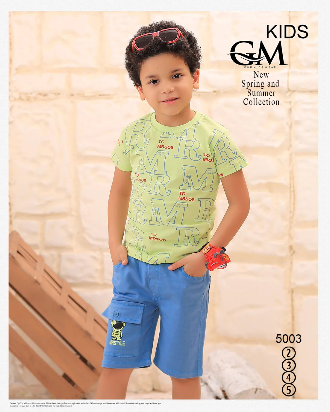 Child wearing a green shirt with text and blue shorts, standing against a light-colored wall.