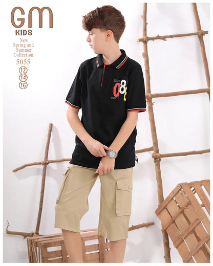 Young boy wearing a black polo shirt with colorful design and beige cargo shorts, standing against a white background with wooden elements.