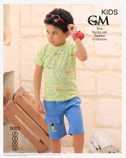 Child wearing a green shirt with text and blue shorts, standing indoors with a neutral background.